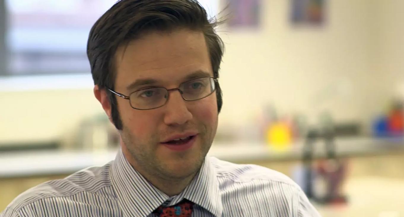 Close up headshot of a young white man with brown hair and glasses wearing a shirt and tie