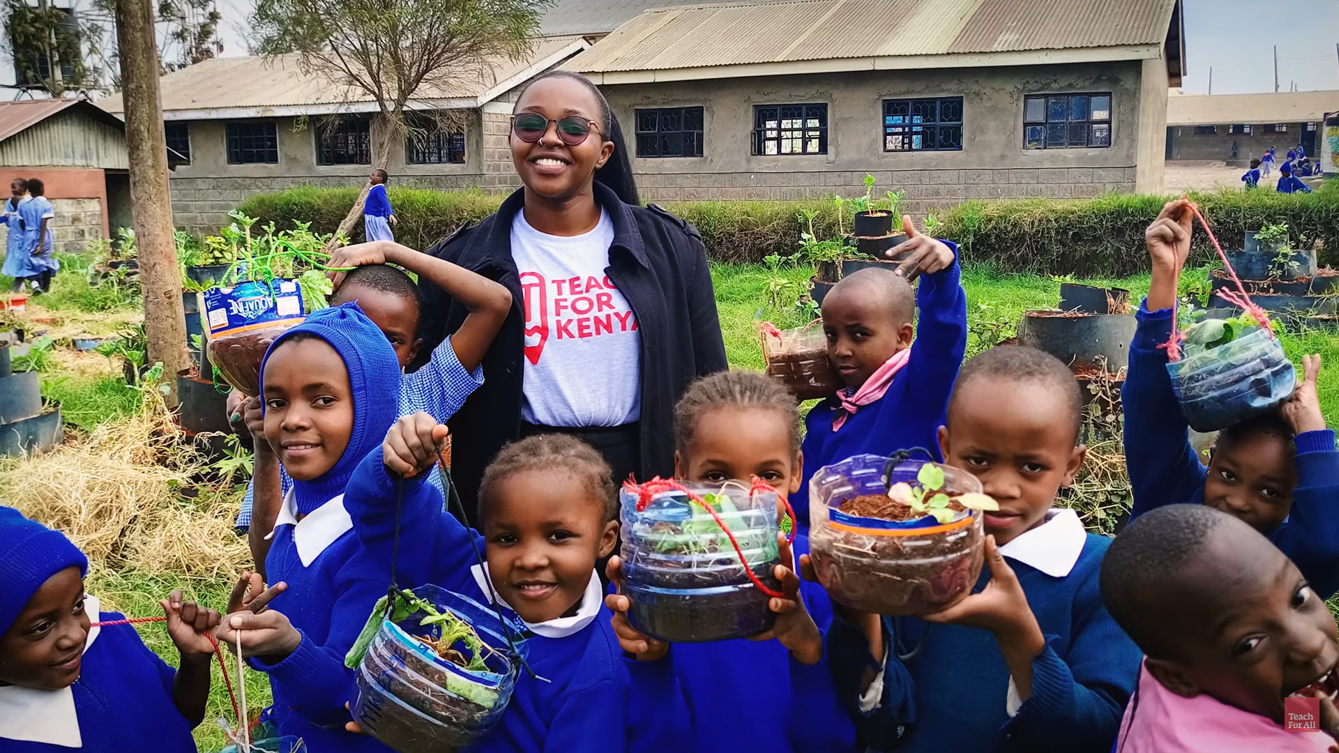 A Black female teachers stands in a school yard surrounded by young students holding up plants growing in recycled plastic containers