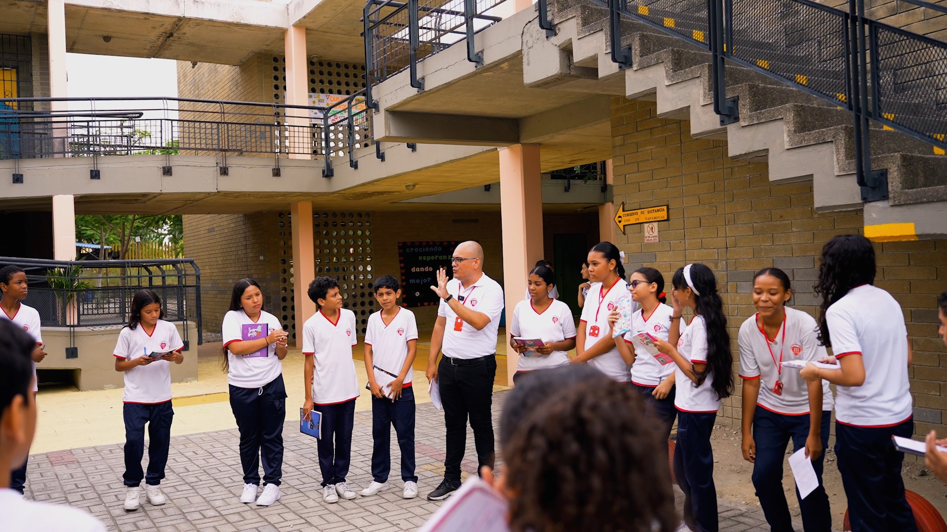A young male teacher in a white shirt with a shaved head stands in a courtyard speaking, surrounded by teenage students also in white shirts