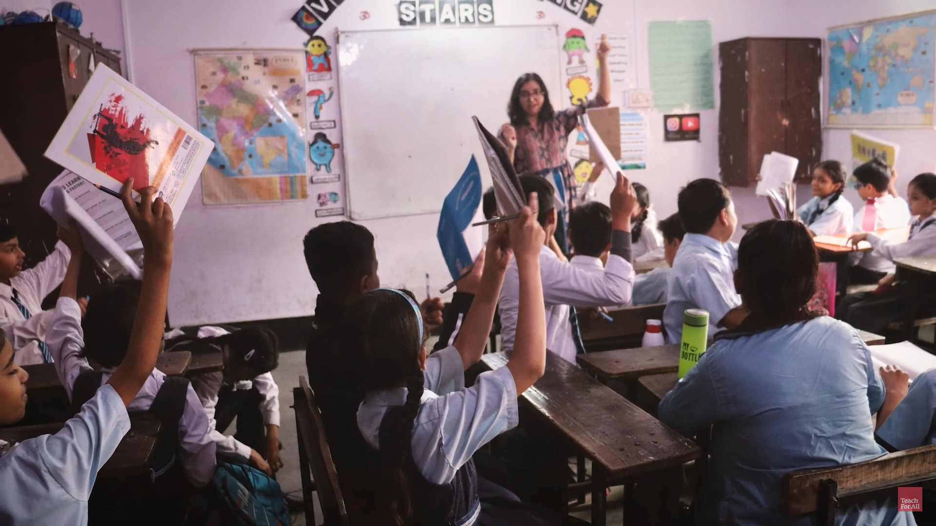 A classroom full of young Indian chilren raise notebooks and smile at their young female teacher standing in the front of a whiteboard