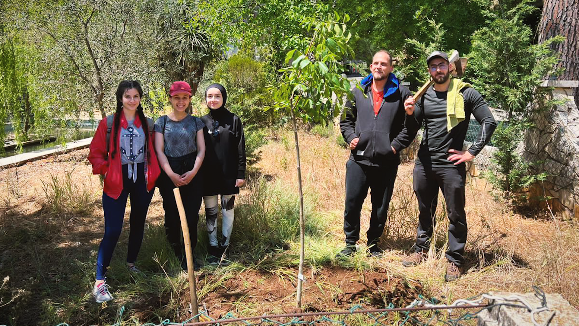 Two bearded men holding shovels and three young teen girls stand near two freshly planted saplings