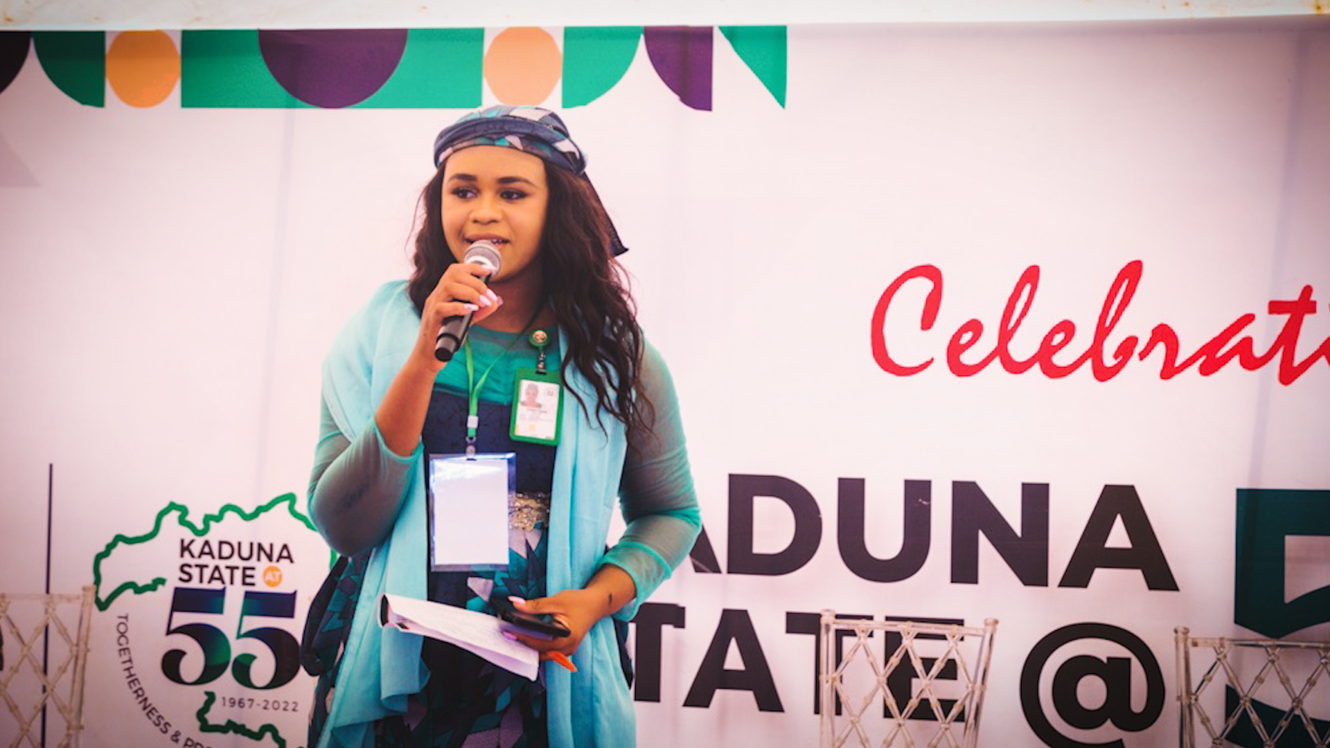 A young light-skinned Black woman with long hair speaks into a microphone on a stage