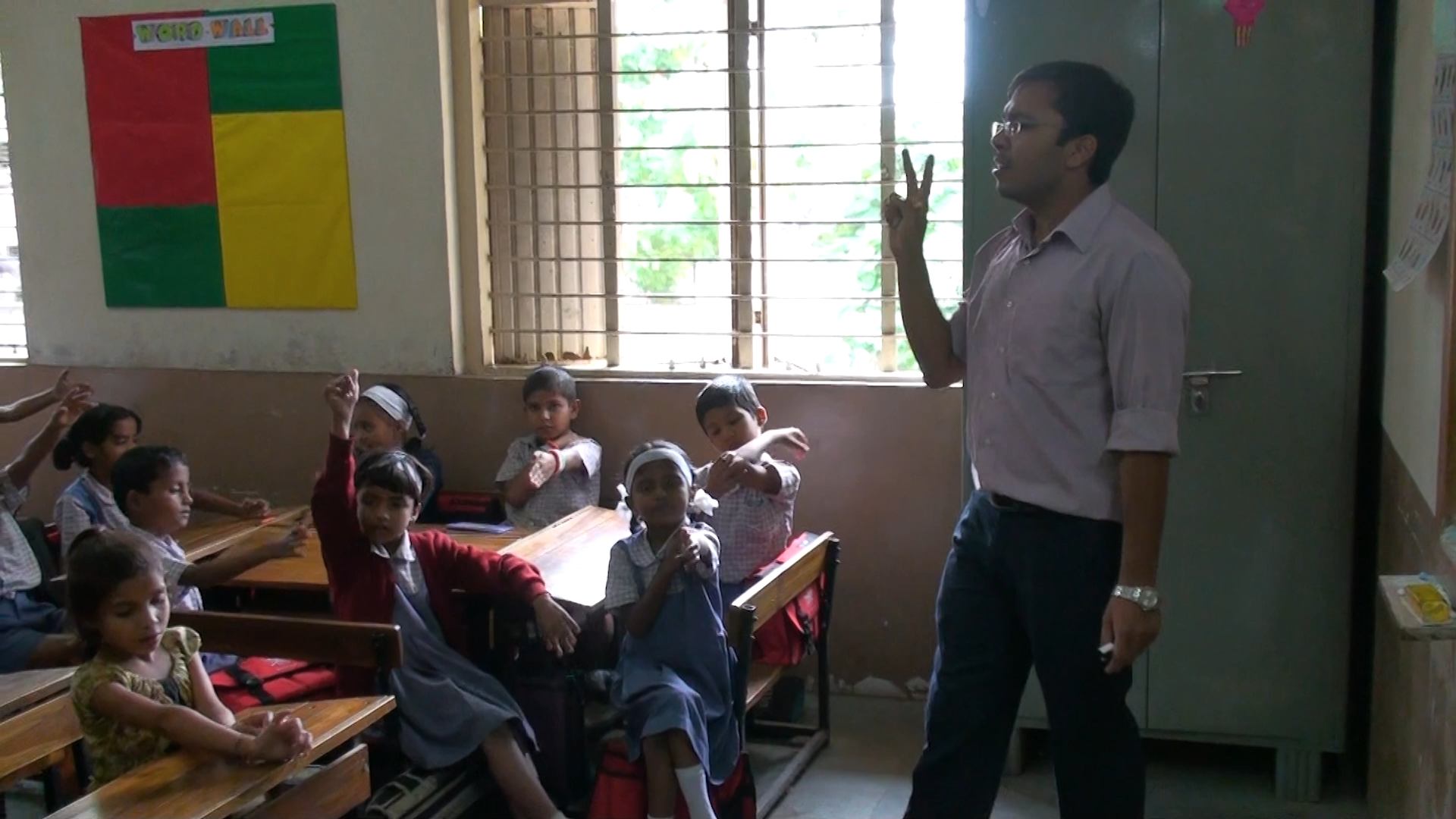 A young Indian male teacher stands in front of his class of young students holding up two fingers