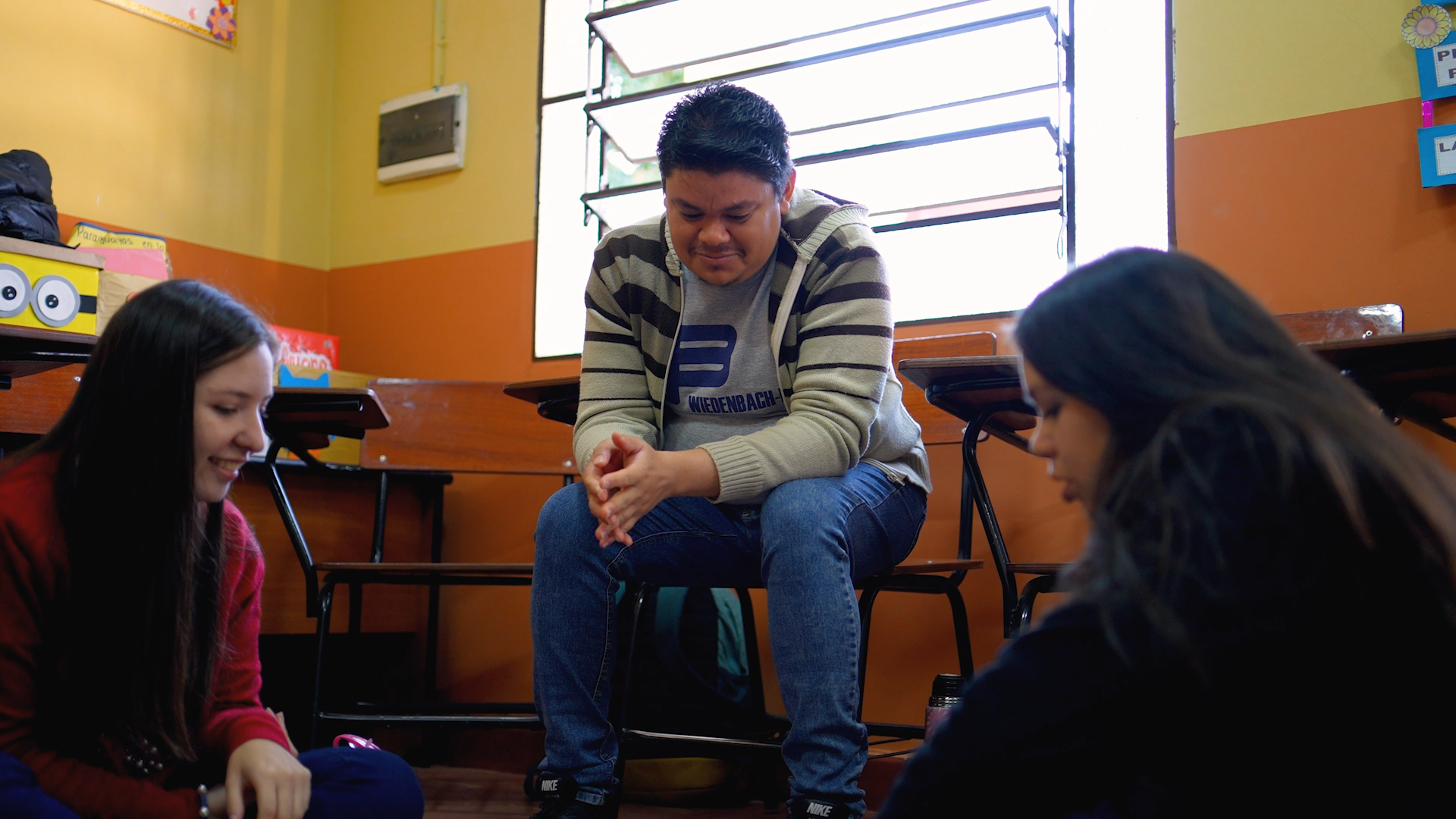 A young man with brown hair sits on a chair smiling while looking at something on the floor, where two young women are seated