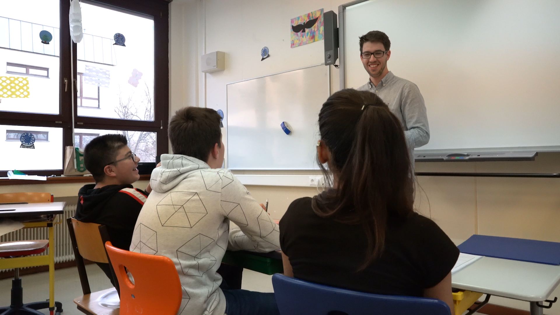 Through the backs of students' heads we see a young white male teacher with brown hair and glasses standing in front of a whiteboard.