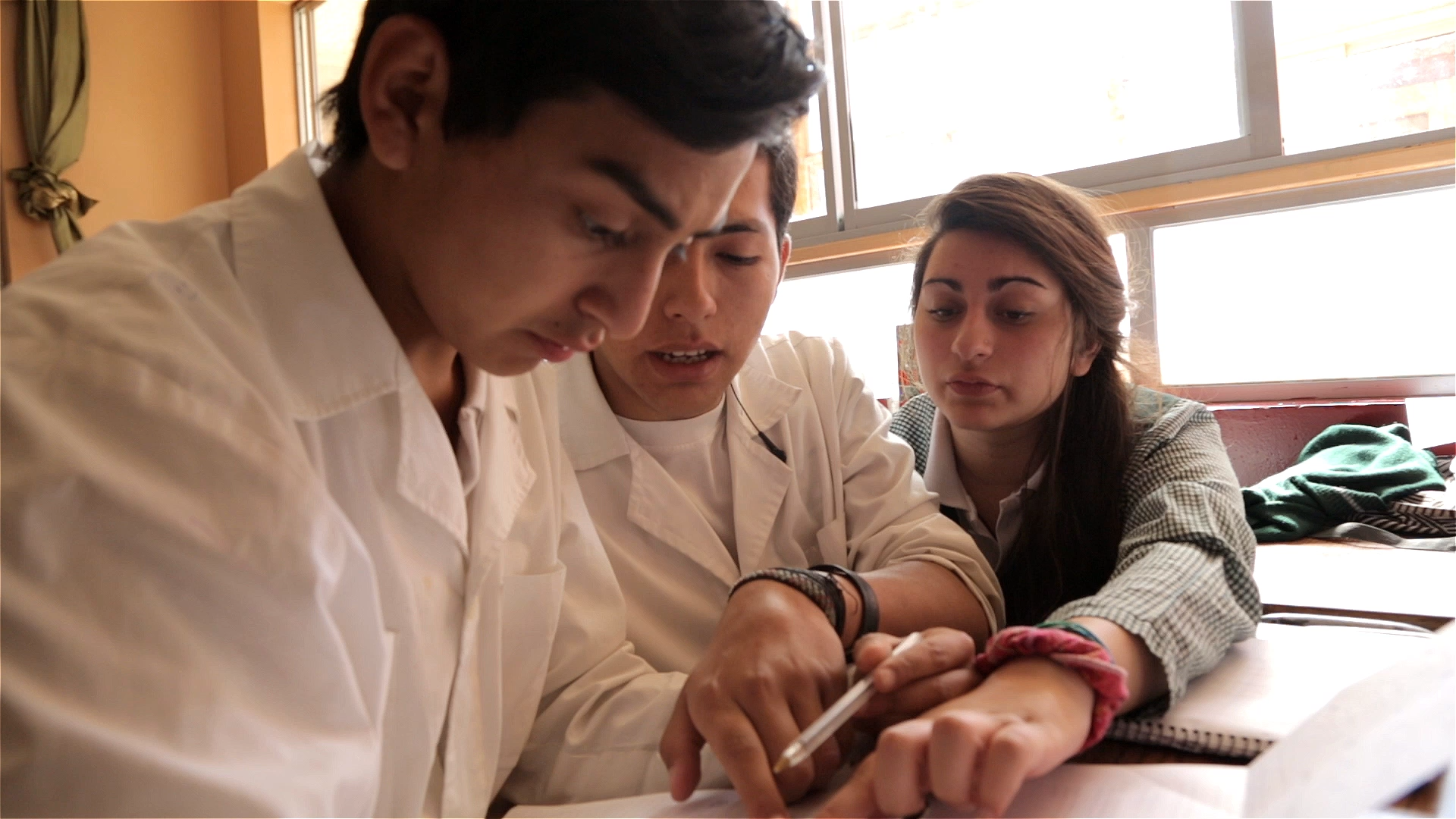 A young woman with long brown hair and two young men with brown hair and tan skin wearing white lab coats work on a problem at a desk