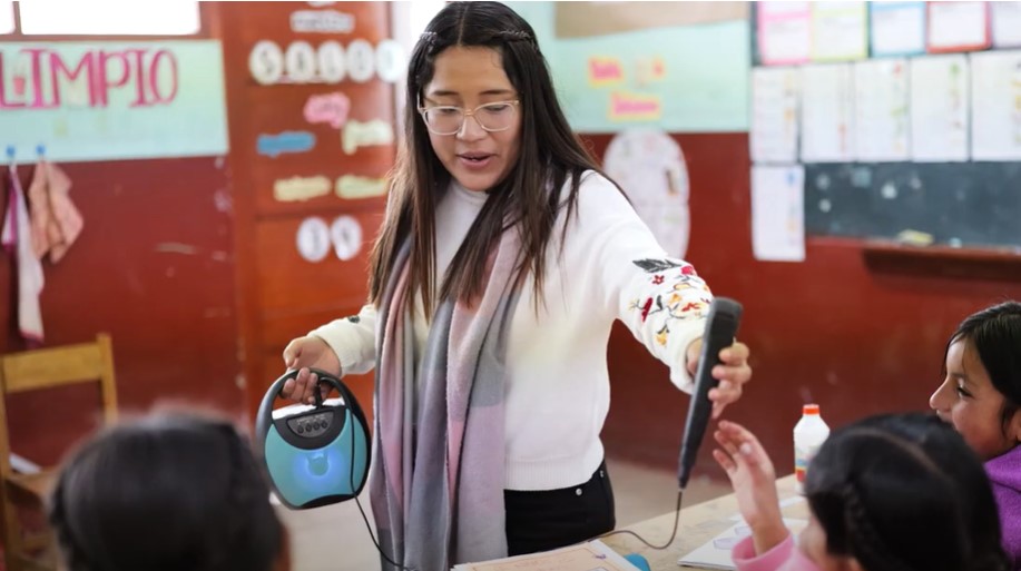 A young woman with long brown hair stands in a classroom speaking to young children