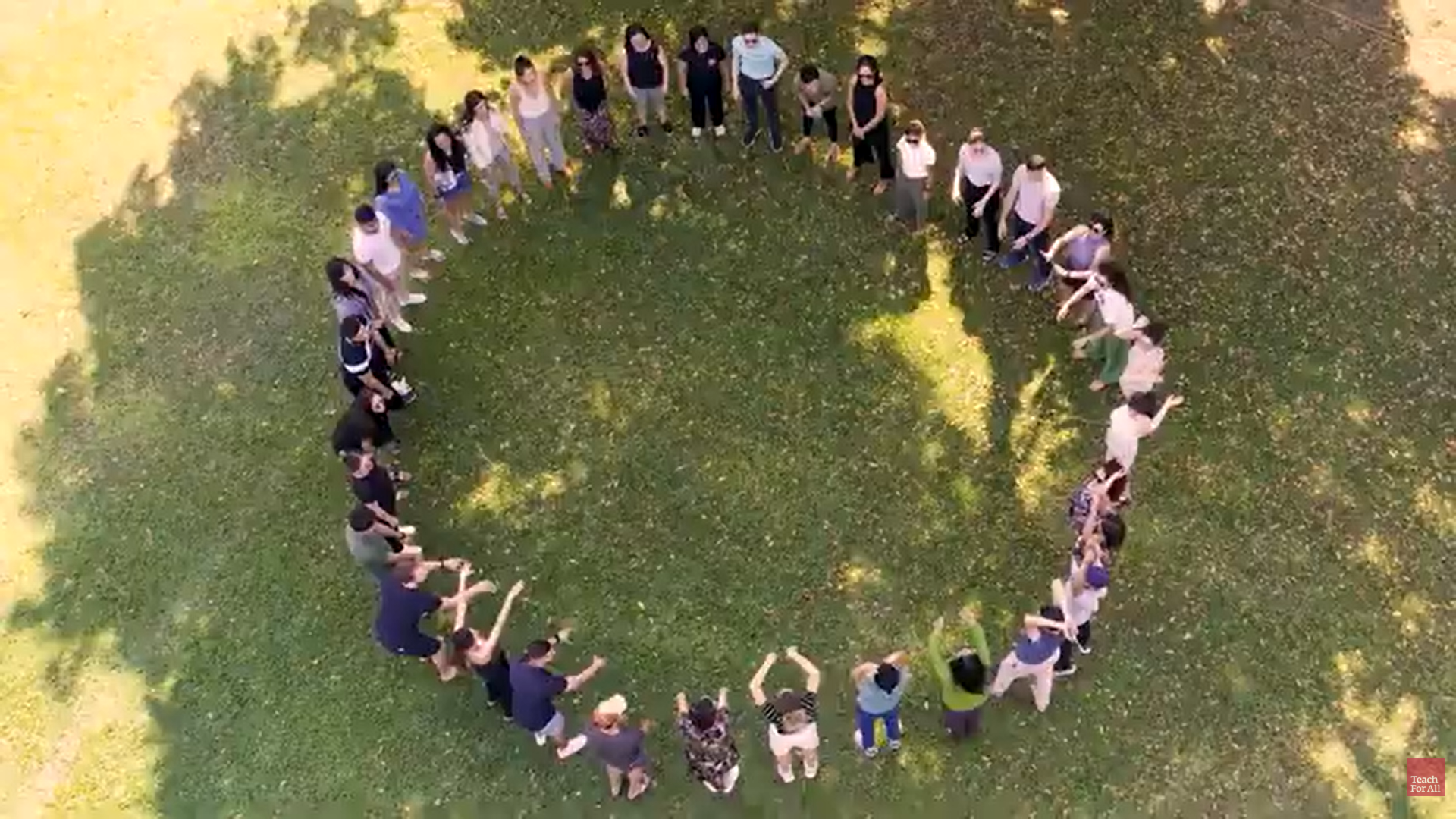 Overhead shot of a group of people standing in a circle on a green lawn with their arms raised