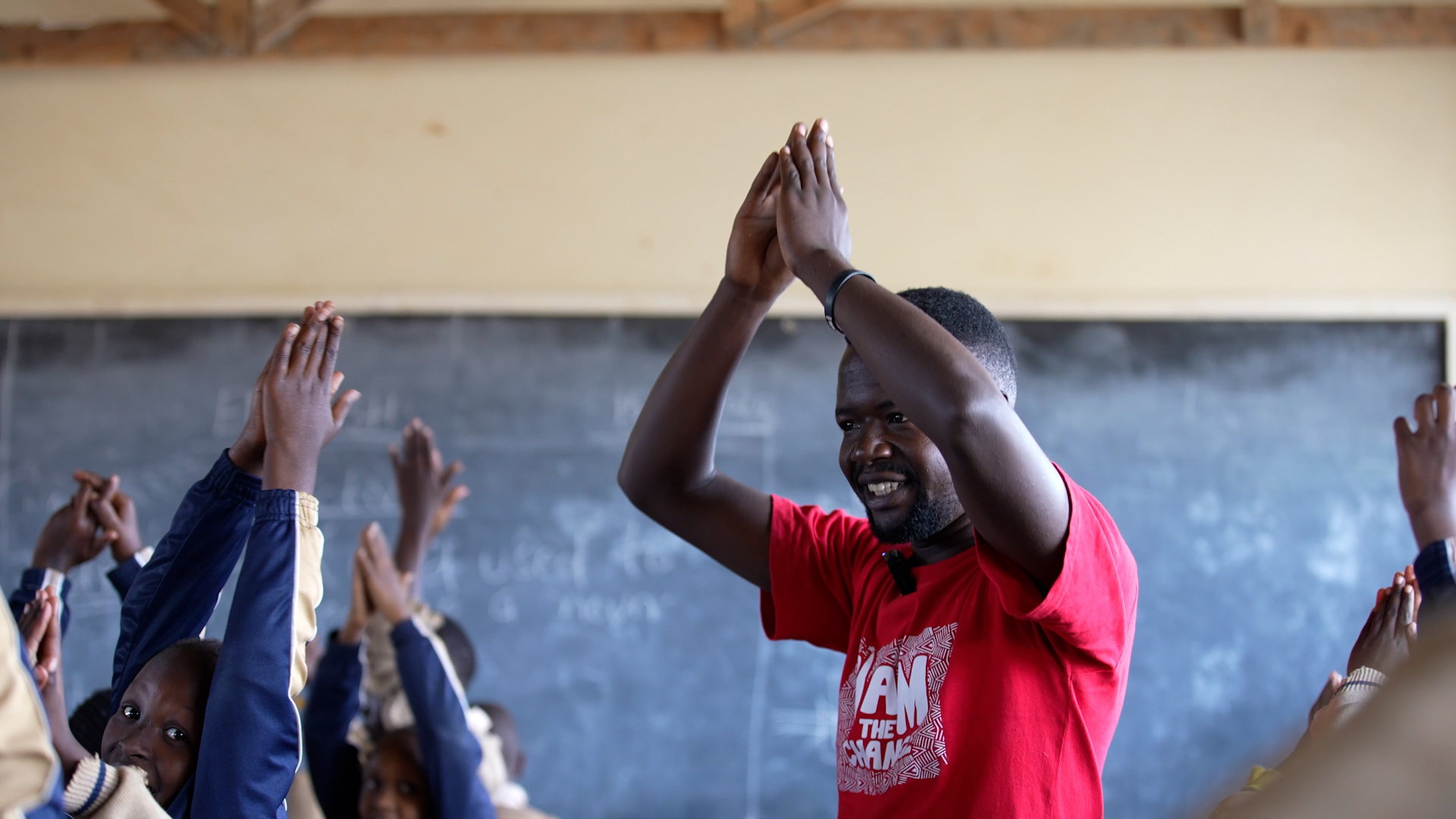 A young Black man claps his hands over his head in a classroom with students doing a similar gesture in front of him
