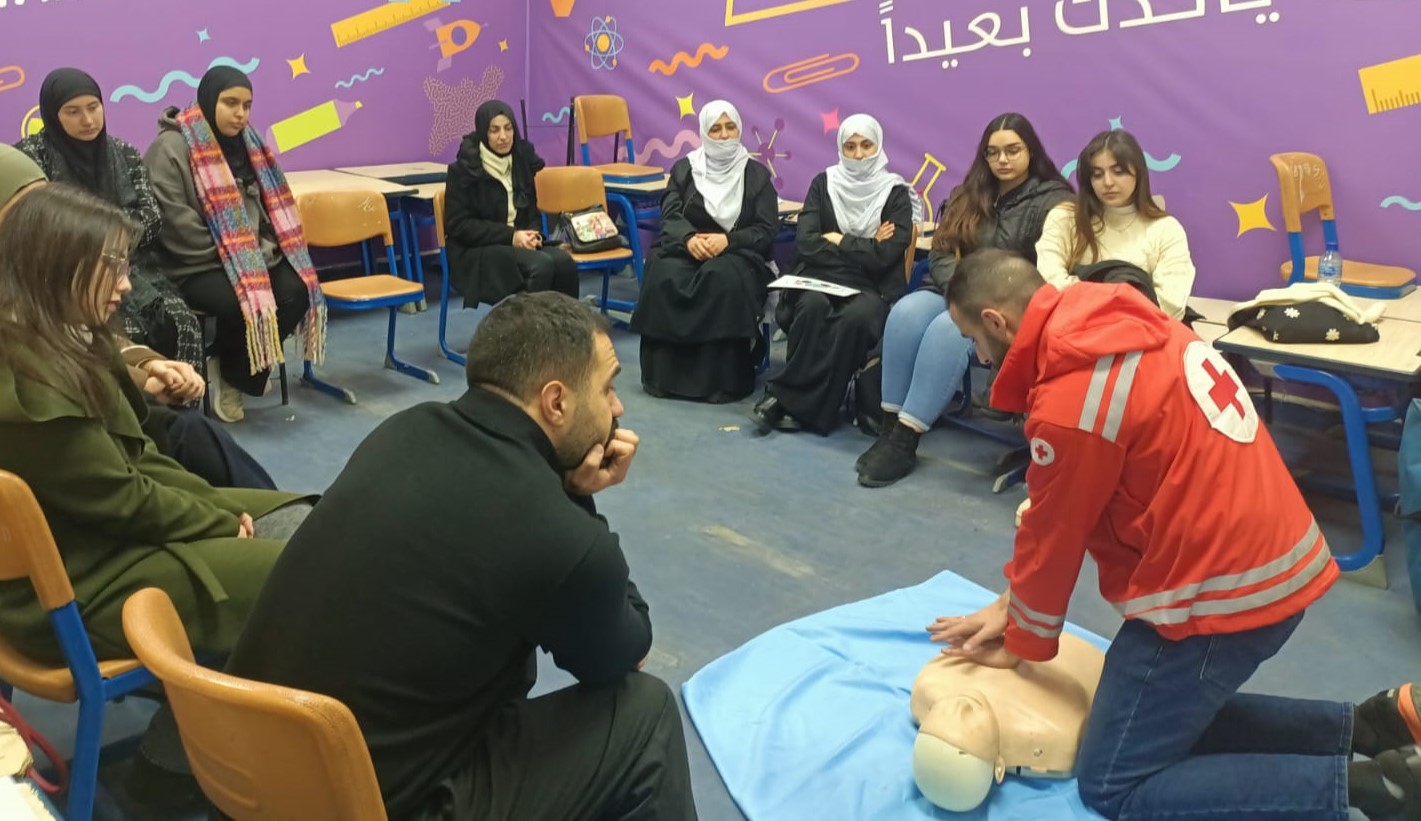 A man in a Red Cross jacket demonstrates CPR on a dummy while a group of people, some in head scarves, watch