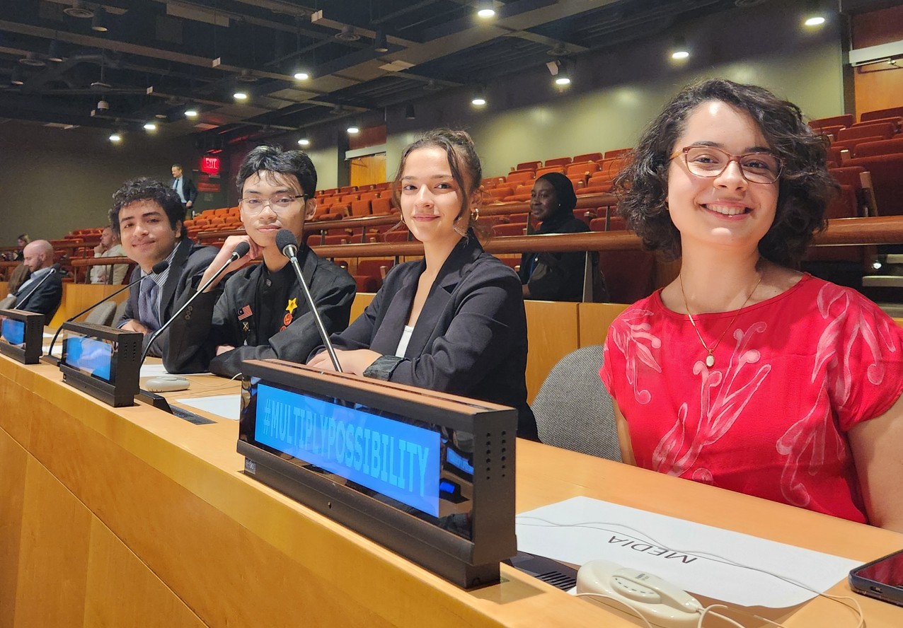 Four young people sit at a panel table equipped with microphones inside a large conference auditorium, behind a digital sign reading "#MULTIPLYPOSSIBILITY"