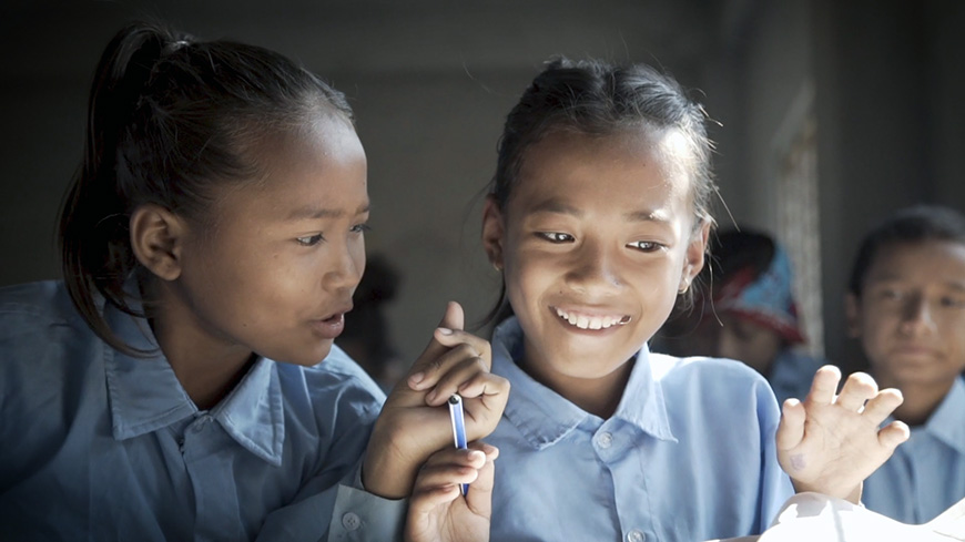 Two Asian girls wearing blue blouses smile while one holds a pen