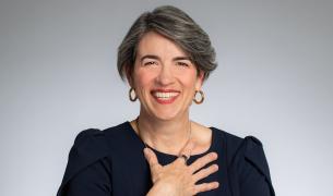 Headshot of a smiling white woman with ear-length black hair touched with grey wearing a black top and pressing her hand to her chest