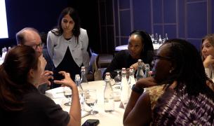 A woman with long brown hair wearing a white blazer leans over a table of diverse adults who appear to be in discussion