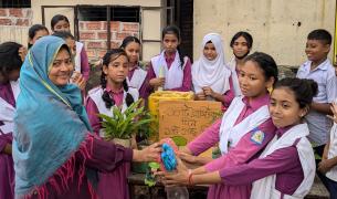A group of South Asian girls in purple school uniforms with white sashes surrounds a project