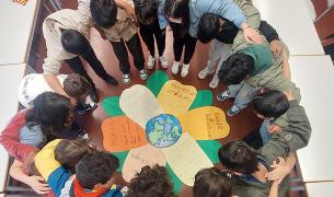 A group of children form a circle with their arms around each other looking down at a paper flower in the middle of the circle