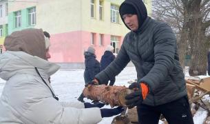 A woman in a black winter jacket and hood hands a log to another person in a light grey winter jacket and hat