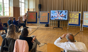 A group of adults sit in chairs in a semi-circle looking at a screen with a presentation and board with post-it notes on them