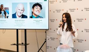 A young woman with light skin and long brown hair holding a microphone addresses an audience next to a screen displaying slide details and speaker headshots for "Education House"