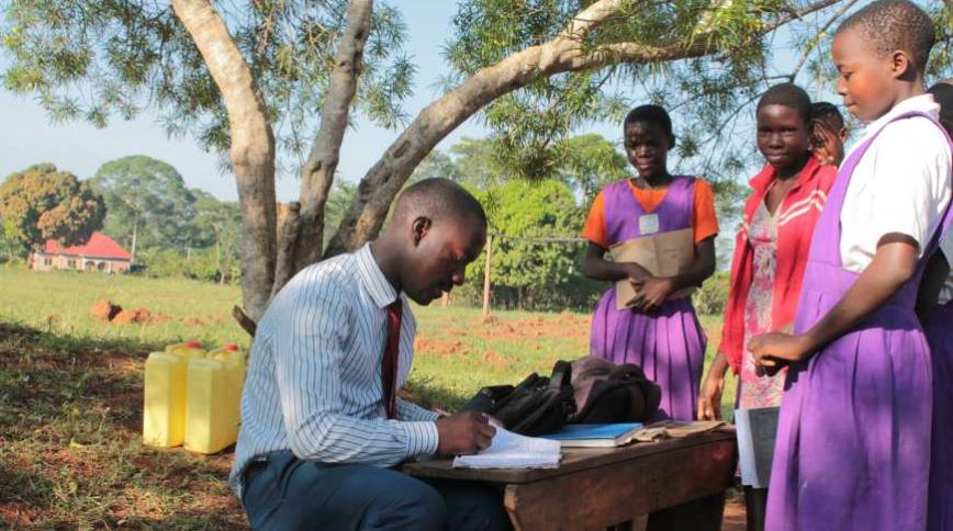Teacher wearing white shirt and red tie sits at outdoor desk with four students looking on.