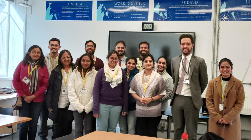 A group of brown-skinned young men and women pose together in front of a classroom
