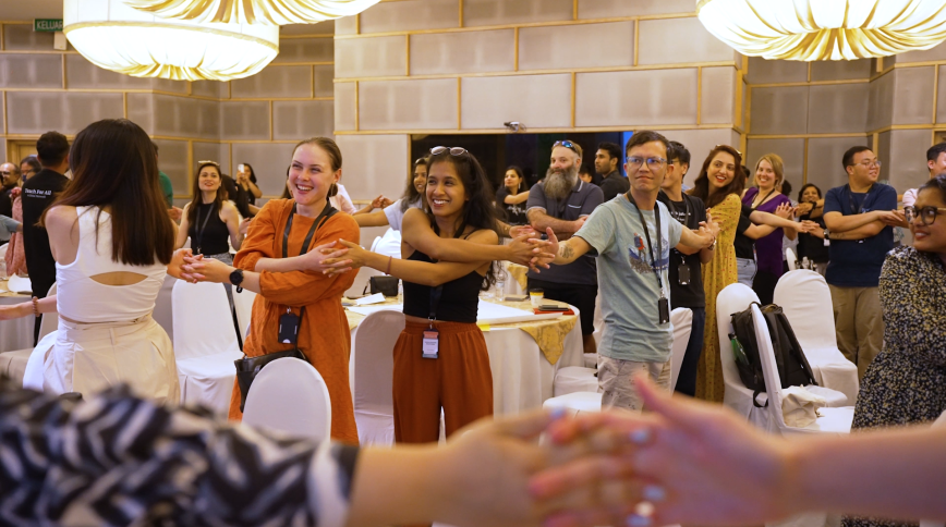 A large group of young adults hold hands while standing in a long chain in a conference room