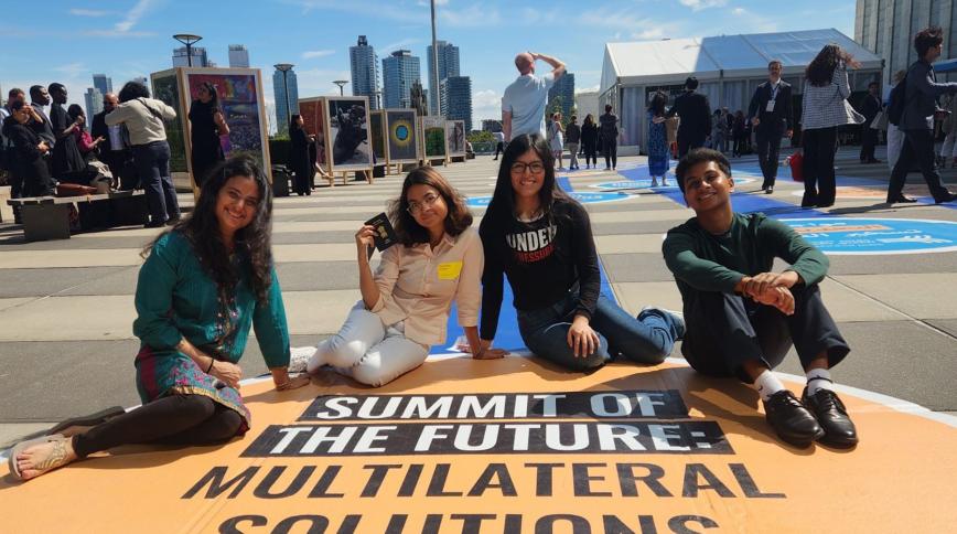 Four diverse young people sit on a yellow street mural that says Summit of the Future Multilateral Solutions with a blue sky and NYC skyline behind them