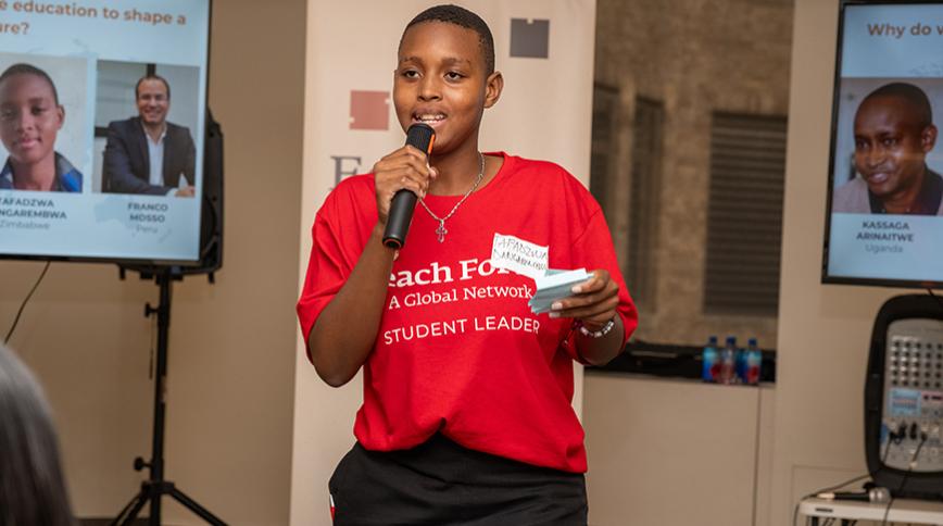 A young african student speaks into a handheld microphone, she is wearing a red shirt that says "Teach For All student leader"