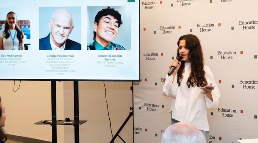 A young woman with light skin and long brown hair holding a microphone addresses an audience next to a screen displaying slide details and speaker headshots for "Education House"