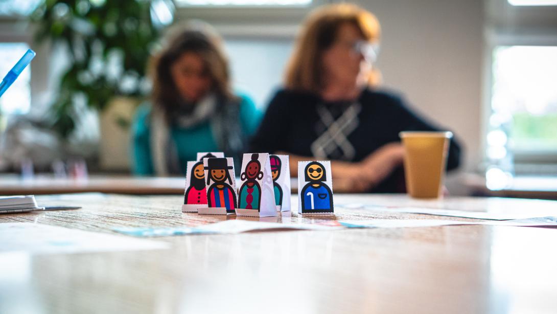 In the foreground are handrawn paper figures of diverse humans on a table while two adult women are visible out of focus in the background seated at another table