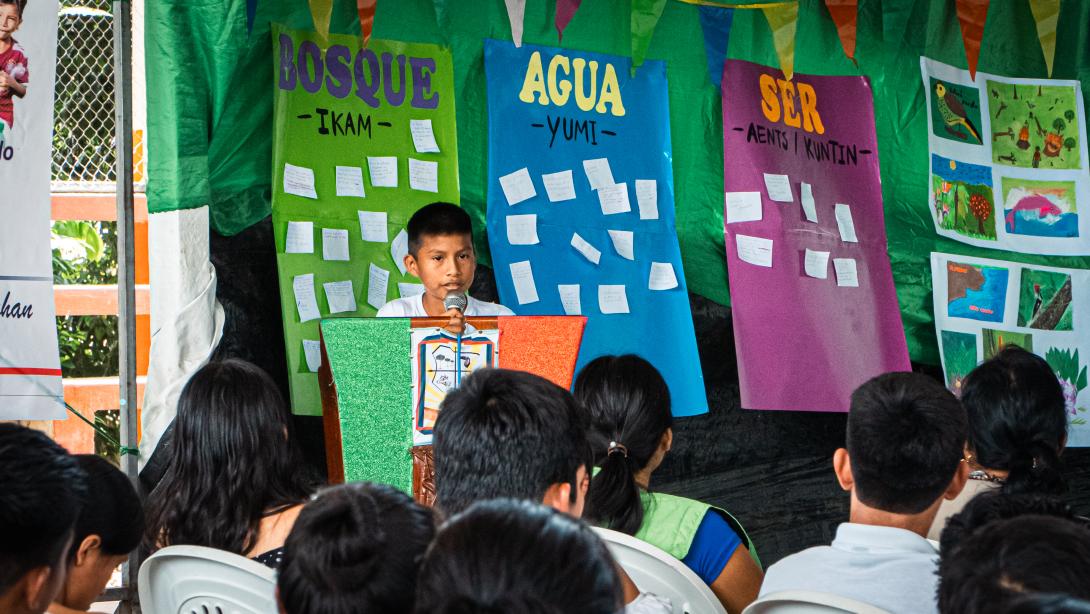 A young boy with tan skin and brown hair speaks at a podium in front of a seated crowd with colorful posters saying "Bosque" "Agua" and "Ser" behind him