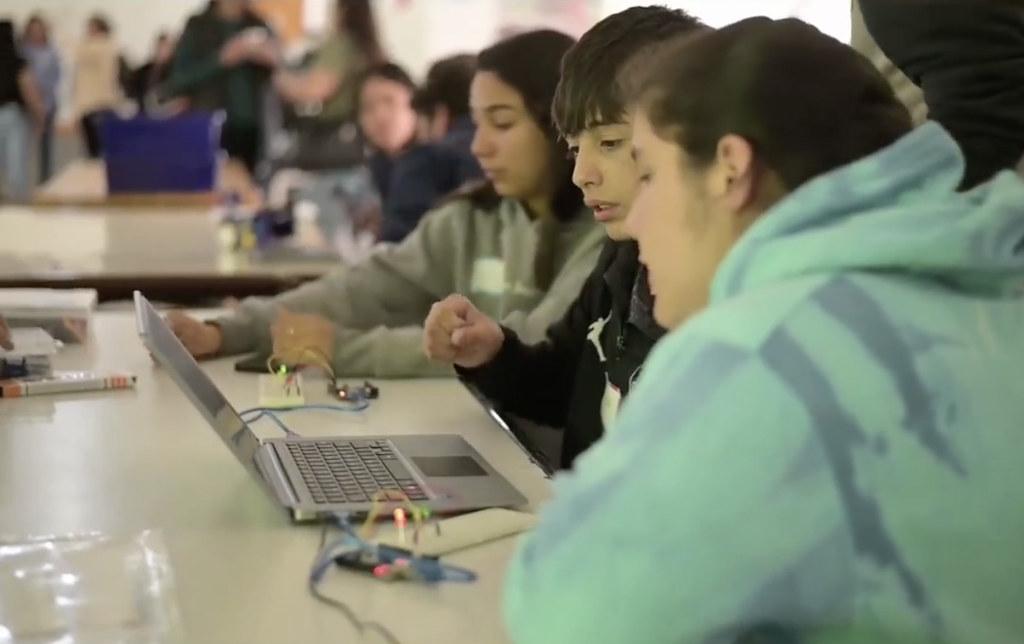 Three teens look at a laptop computer at a table, the girl closest to the camera wears a tie-dyed green sweatshirt.