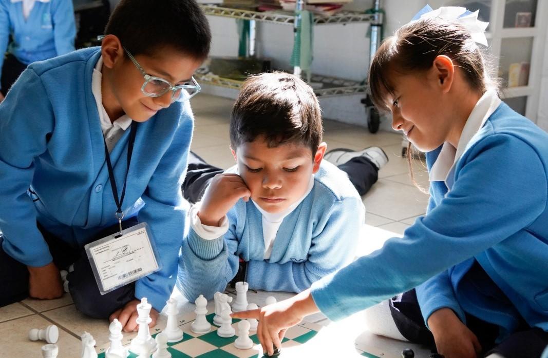 Two young boys and a young girl with brown hair and tan skin in blue school uniforms play chess on the floor