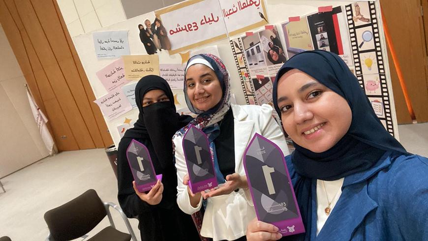 Three women in hijabs pose holding first place trophies in front of a poster with Arabic text and photos on it