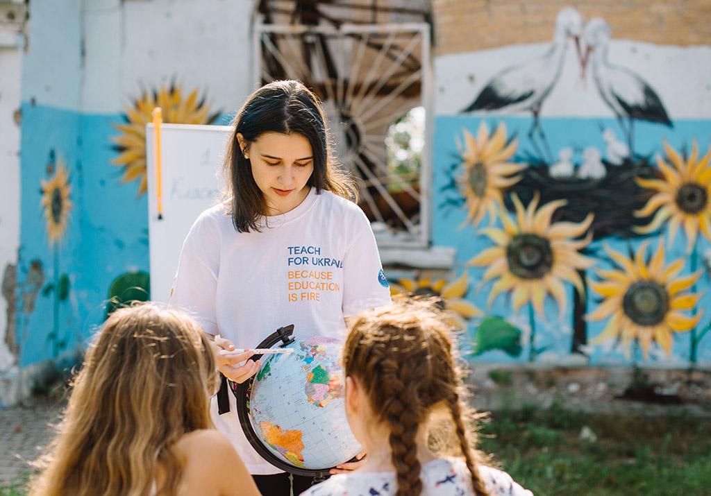 A female teacher with long brown hair shows a globe to two female students we see from behind, with a backdrop of outdoor walls painted with sunflowers