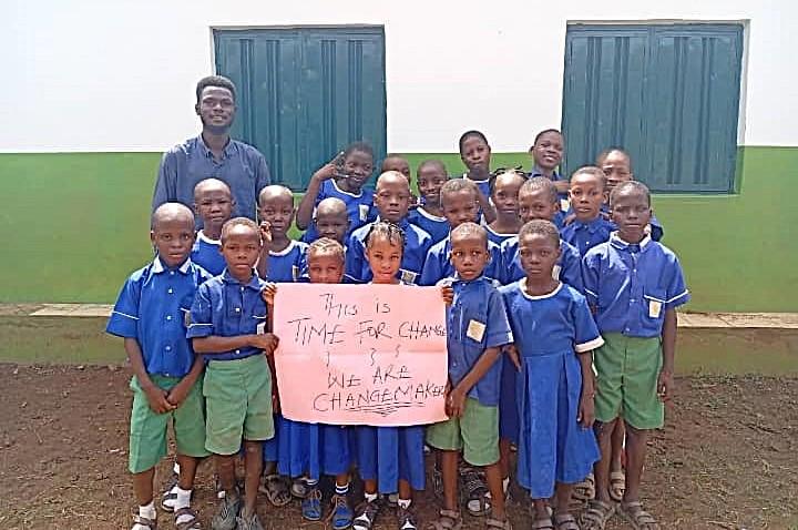 A group of African children in blue and green school uniforms and their male teacher stand together holding a sign that reads "This is Time for Change. We are Changemakers"