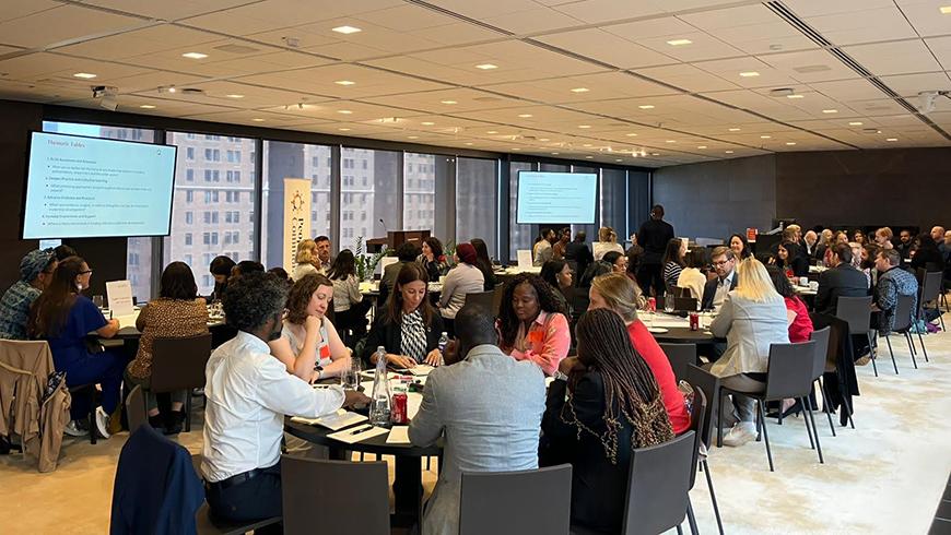 A wide image of a meeting room filled with people in business casual clothing sitting at round tables