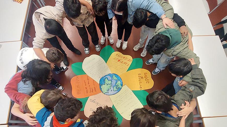 A group of children form a circle with their arms around each other looking down at a paper flower in the middle of the circle